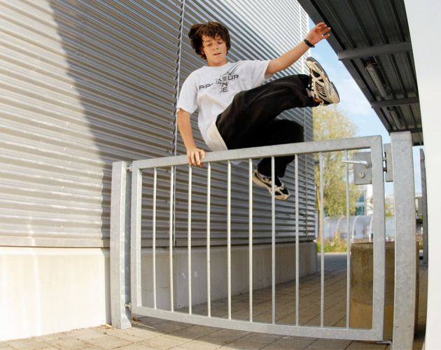 Parkour: Die Kunst der effizienten Fortbewegung Ein Jugendlicher hält sich an einem Gitter und schwingt sich mit Beinen voran darüber.