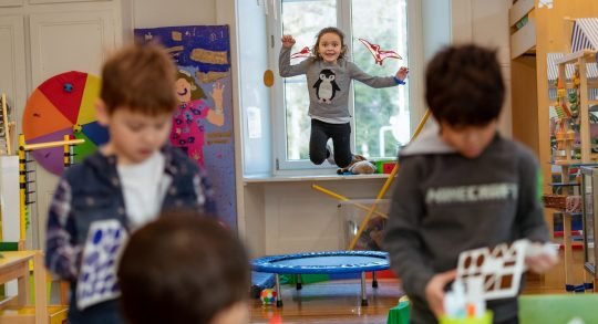 Disegno: tre bambini in primo piano eseguono dei compiti cognitivi mentre una bambina dietro di loro salta sul minitramp