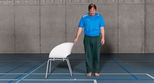 Mann dehnt stehend die Nackenmuskulatur und hält sich an einem Stuhl.