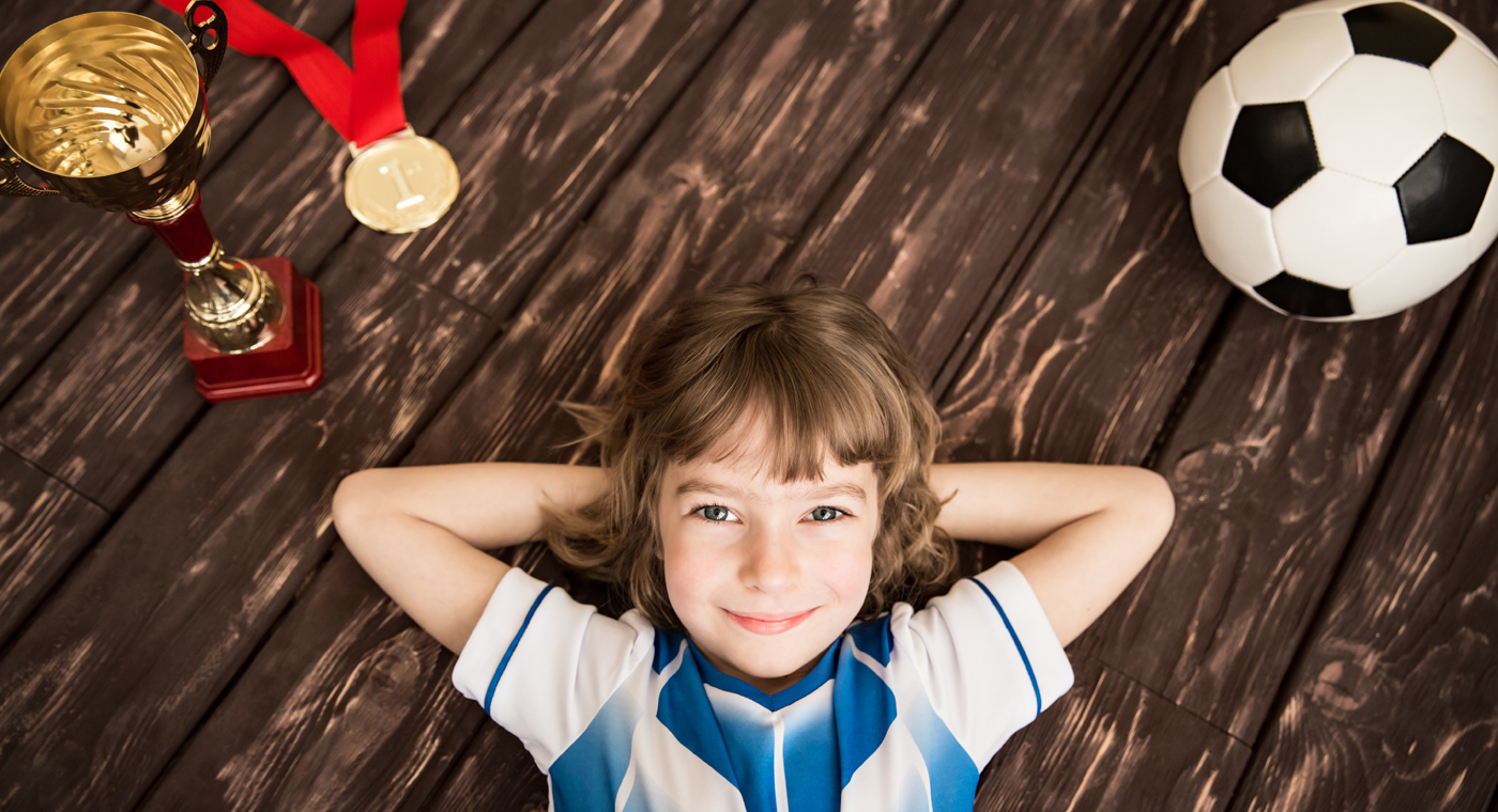 Lächelndes Mädchen liegt zufrieden am Boden zwischen Pokal, Medaille und Fussball.